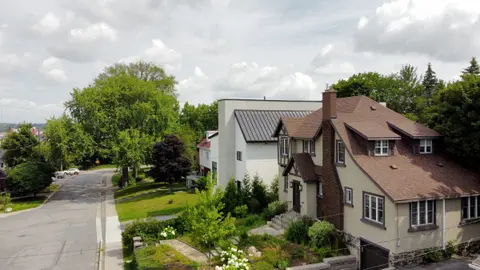 The finished house, white with a dark metal roof, on its narrow lot beside a Tudor-style neighbour
